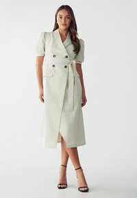 Vestido de color beige claro, de longitud hasta la rodilla, con un diseño de doble botonadura, mangas cortas abullonadas, cinturilla con lazo y abertura frontal; cuenta con detalles de botones y bolsillos laterales.