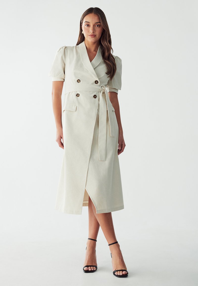 Vestido de color beige claro, de longitud hasta la rodilla, con un diseño de doble botonadura, mangas cortas abullonadas, cinturilla con lazo y abertura frontal; cuenta con detalles de botones y bolsillos laterales.