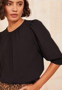 Blusa nera con maniche corte a sbuffo, scollatura plissettata e polsini texturizzati. Indossata con una gonne a fantasia. Sfondo chiaro.