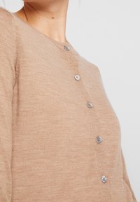 Beige svetr s kulatým výstřihem, se pěti stříbrnými knoflíky vpředu, vyrobený z texturované pleteniny.