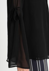 Svart genomskinlig blus med breda ärmar som knyts vid handleden. Tyget verkar lätt och flödande, med en slät yta.