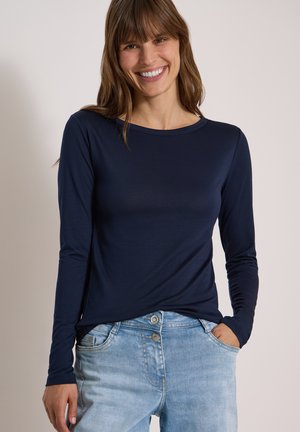Femme souriante aux cheveux bruns portant une chemise bleu marine à manches longues et un jean bleu clair, une main dans la poche.