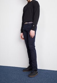Schwarzes Langarmhemd, dunkelblaue Hose und schwarze Sneaker mit braunen Sohlen. Hemd hat enge Manschetten; der Gürtel ist grau. Steht an einer weißen Wand.