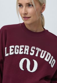 Burgunderfarget hettegenser med hevet hvit tekst som sier "LEGER STUDIO" og et dekorativt løkkemønster. Ribbestrikket hals og teksturert stoff.