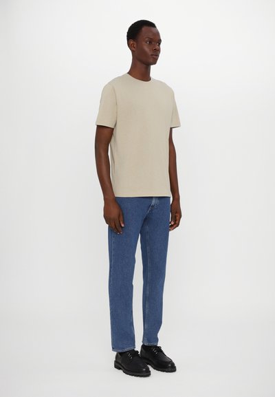 Camiseta de algodón beige de manga corta, combinada con jeans azul de pierna recta y zapatos negros de cordones. El modelo se encuentra frente a un fondo neutro.