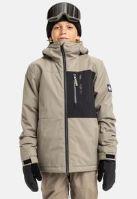 Chłopiec w beżowej i czarnej zimowej kurtce, beżowych spodniach, czarnych rękawiczkach, czapce beanie w kolorze czarnym i gogle narciarskie na czole, na białym tle.