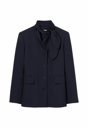 Femme portant un blazer bleu marine foncé avec une ceinture marron, un pantalon ample couleur crème, des chaussures marron et une écharpe nouée autour du cou, debout sur un fond uni.