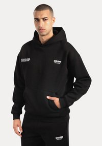 Czarny hoodie wykonany z miękkiego materiału, z kieszenią kangurką i prążkowanymi mankietami. Biały napis z logo na klatce piersiowej i rękawie. Prosty, luźny krój.