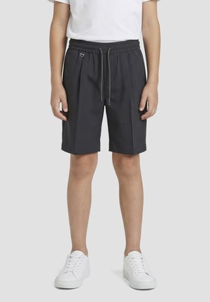 Personne portant un short noir à taille élastique avec des cordons, associé à des baskets blanches et un t-shirt blanc, debout devant un fond uni.