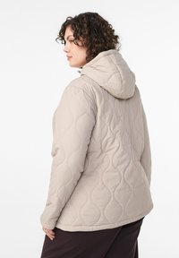 Beige quiltet jakke med hætte, der har en blød tekstur og buede mønsterdetaljer. Designet giver en afslappet pasform for komfort.