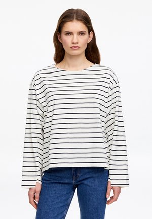 Kvinde med langt brunt hår iført en hvid langærmet skjorte med tynde sorte, horisontale striber og blå jeans, stående med fronten mod kameraet.