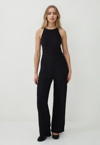 Vrouw met lang blond haar, gekleed in een mouwloze zwarte top, gestructureerde zwarte wijde broek en zwarte hakken, staand op een witte vloer.