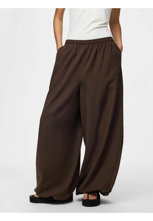 Person iført mørkebrune, brede bukser med elastik i taljen og sorte plateausandaler, stående med hænderne i lommerne.