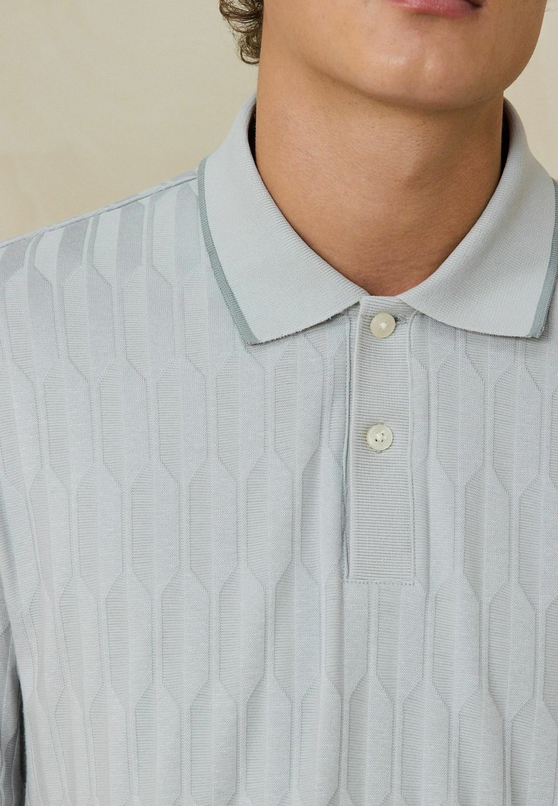 Gros plan d'une personne portant un polo gris clair texturé avec un col et deux boutons avant attachés.