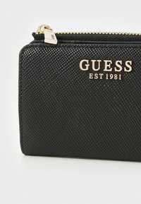 Portafoglio nero con texture e chiusura a zip, con un dettaglio in oro che riporta la scritta "GUESS EST 1981". Materiale sintetico liscio.