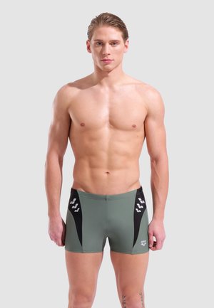 Gespiereerde jonge man met nat haar die groene en zwarte aansluitende zwemshorts draagt, staand tegen een effen lichte achtergrond.