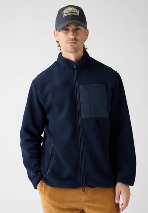 Man draagt een navy fleecejas met ritssluitingen en een borstzak, gecombineerd met een blauwe pet met de tekst "Knowledge" en bruine corduroy broek.