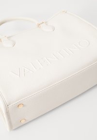 Vit texturerad läderväska med prägling av "Valentino"-logotyp, detaljer i guldton och nitar på botten för hållbarhet.