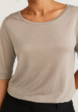 Femme portant un haut beige à manches courtes et encolure dégagée, rentré dans un pantalon noir, sur un fond uni.