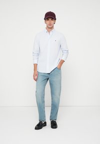 Ανοιχτό μπλε πουκαμίσου με ρίγες, χαλαρές μπλε τζιν και μαύρα μοκασίνια. Η βουργουνδί καπέλο ολοκληρώνει την εμφάνιση, όρθιο σε λευκό φόντο.