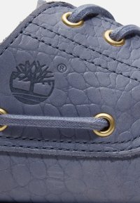 Κοντινό πλάνο μπλε παπούτσι από δέρμα με υφή, με μεταλλικά κουμπώματα και κορδόνια, που διαθέτει ανάγλυφο λογότυπο δέντρου Timberland και σύμβολο κατατεθειμένου εμπορικού σήματος.