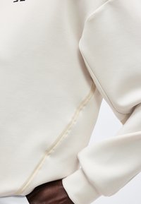 Nærbilde av en person som har på seg et kremfarget plagg med lange ermer og synlige sømdetaljer langs siden og håndleddet.