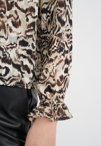 Blusa con stampa leopardata in tessuto trasparente, con dettagli di maniche a sbuffo. La blusa contrasta con la pelle nera testurizzata sottostante.