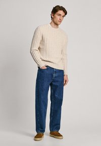 Cremfarbener Zopfmuster-Pullover mit gerippter Textur, kombiniert mit dunkelblauen Jeans. Das Model trägt braune Wildlederschuhe und steht vor einem neutralen Hintergrund.