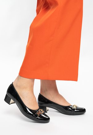 Schwarze Pumps aus Lackleder mit niedrigem Absatz, verziert mit einem goldfarbenen Akzent und einer abgerundeten Spitze. Getragen zu einem orangefarbenen Kleidungsstück.