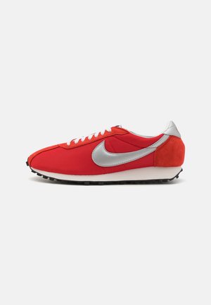 Czerwona niska tenisówka z białymi sznurowadłami, srebrnym logo Nike, zamszową łatką na pięcie i czarną podeszwą wafelową na białym tle.