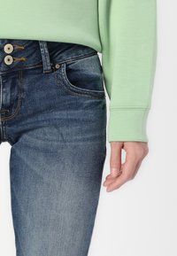 Jean en denim bleu ajusté avec un délavage, design à cinq poches et détails de couture visibles ; associé à un haut à manches longues vert menthe.