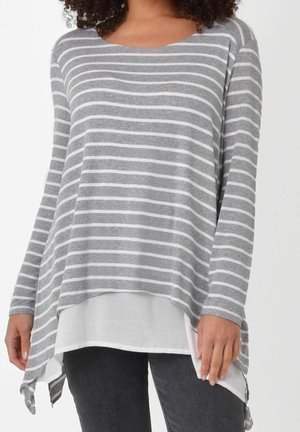 Femme portant une tunique à manches longues rayée gris et blanc avec un ourlet en pointe au-dessus d’un chemisier blanc et d’un pantalon foncé.