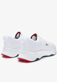 Baskets blanches de sport avec une tige en mesh, semelle blanche texturée, accents rouge et marine au niveau du talon et de la languette, et un design rembourré et offrant un bon soutien.