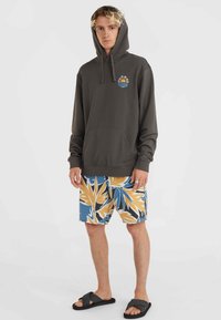 Donkergrijze hoodie met een voorzak en een kleurrijke zon- en golfafbeelding. Gecombineerd met multikleurige bloemen zwemshorts en zwarte sandalen.