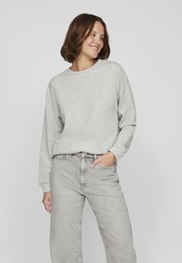 Mujer joven con pelo corto castaño, vistiendo un sudadera gris claro y jeans de talle alto gris deslavado, sonriendo y mirando de lado.