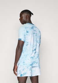 Junger Mann mit geflochtenem Haar, der seitlich vor einem schlichten Hintergrund steht, und ein passendes hellblau marmoriertes Hemd und Shorts trägt.