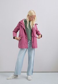 Chaqueta impermeable rosa con capucha, cierre de botones en la parte delantera y dos bolsillos laterales. Llevada sobre un suéter verde y combinada con unos pantalones vaqueros de pierna ancha de color azul claro.