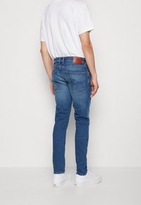 Blå denimjeans med slim fit, fem fickor och en brun läderlapp på baksidan av midjan. Kommer med vita sneakers.