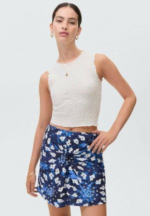 Witte cropped top van gestructureerde stof met geschulpte randen, gecombineerd met een blauwe bloemenrok met een wikkelontwerp, voorzien van verschillende tinten blauw en witte patronen.