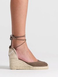 Jambe féminine portant une espadrille compensée marron avec lacets à nouer autour de la cheville et semelle plateforme en jute tressée sur fond gris clair.