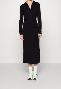Robe noire à manches longues avec col chemise, taille cintrée à l'aide d'un lien, de longueur midi ; portée avec des chaussettes blanches à la cheville et des baskets vertes en accent.
