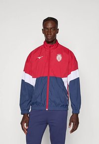 Rød, hvid og marineblå vindjakke med elastiske manchetter og taljebånd. Har et logo på brystet og fuld lynlås foran.