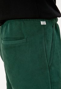 Joggers en coton vert avec une ceinture élastique, dotés d'une poche latérale et d'une étiquette "Q/S". Texture lisse et design décontracté.