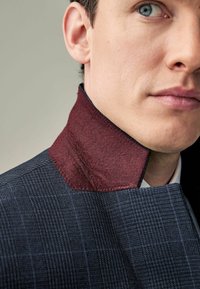 Blau gemusterter Stoff mit einem strukturierten, kastanienbraunen Kragen. Der Kragen hat eine kontrastierende Naht und ist Teil eines maßgeschneiderten Kleidungsdesigns.