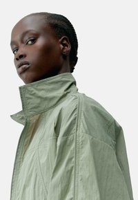 Světle zelená bunda s vysokým límcem, vyrobená z texturované látky, s čistými liniemi a minimálními designovými detaily. Slim fit a moderní silueta.