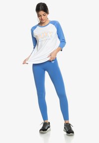 Roxy träningsset bestående av en vit och blå tröja med trekvartsärmar och logotyp samt blå ribbade leggings, tillsammans med svarta sneakers.