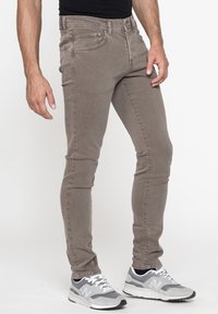 Jeans in denim marrone slim-fit con una texture liscia, design a cinque tasche e passanti per cintura, abbinati a sneakers athletic grigie.