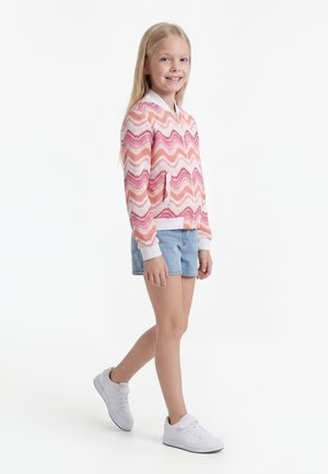 Fille souriante marchant, portant une veste à motif zigzag rose et orange, un short en jean bleu clair et des baskets blanches sur fond blanc.