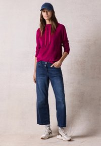Felpa cappuccio fucsia con coulisse, abbinata a jeans a gamba larga blu. Il modello indossa un cappellino blu navy e sneakers bianche con dettagli grigi.