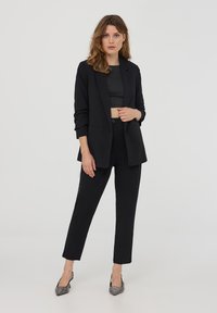 Blazer nero su misura con vestibilità rilassata, top nero corto abbinato e pantaloni neri slim-fit. Tacchi con dettagli argentati completano il look.